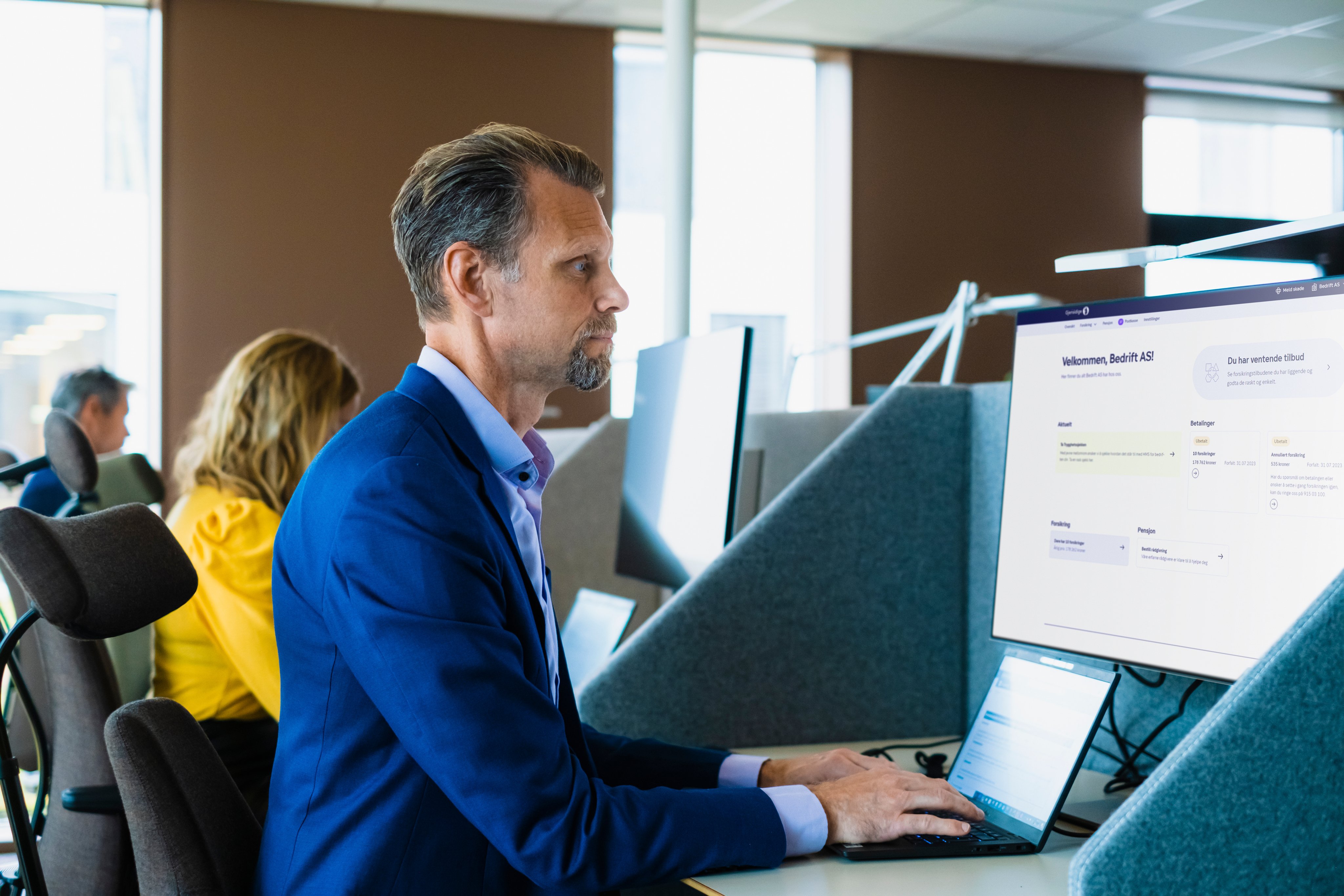 Mann sitter foran to pc-skjermer i et kontorlandskap.