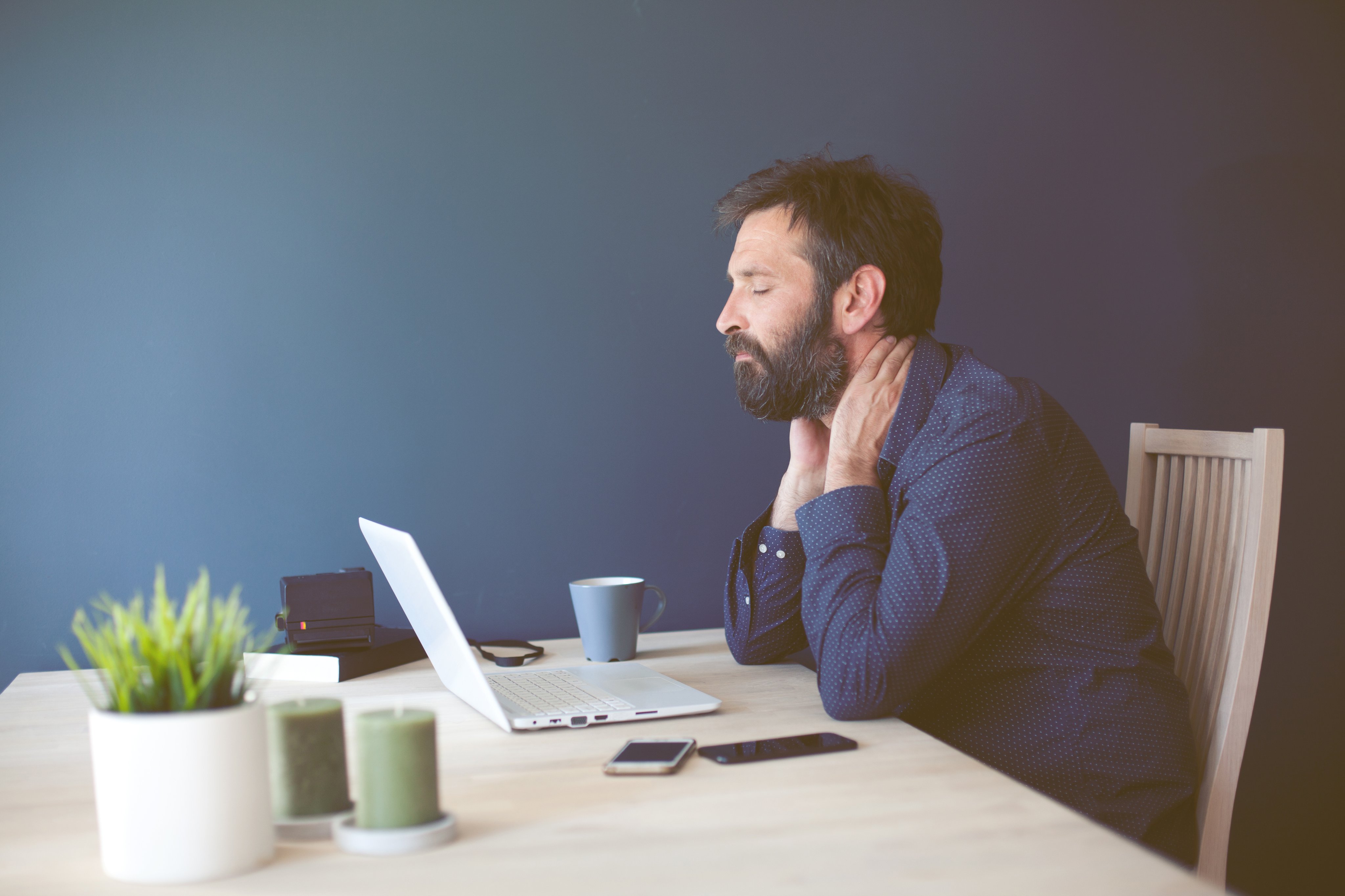 En mann som sitter ved en laptop og masserer nakken som om han har vondt.