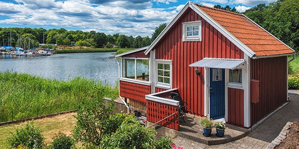 Rødt trehus ved vannkanten med grønn vegetasjon og blå himmel.