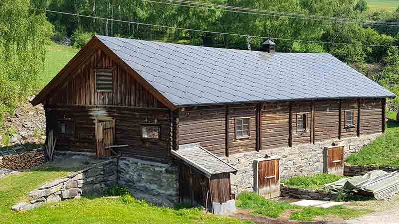 Lang, tradisjonell trebygning med steinfundament og mørkt skråtak, omgitt av grønn natur og trær på landsbygda.
