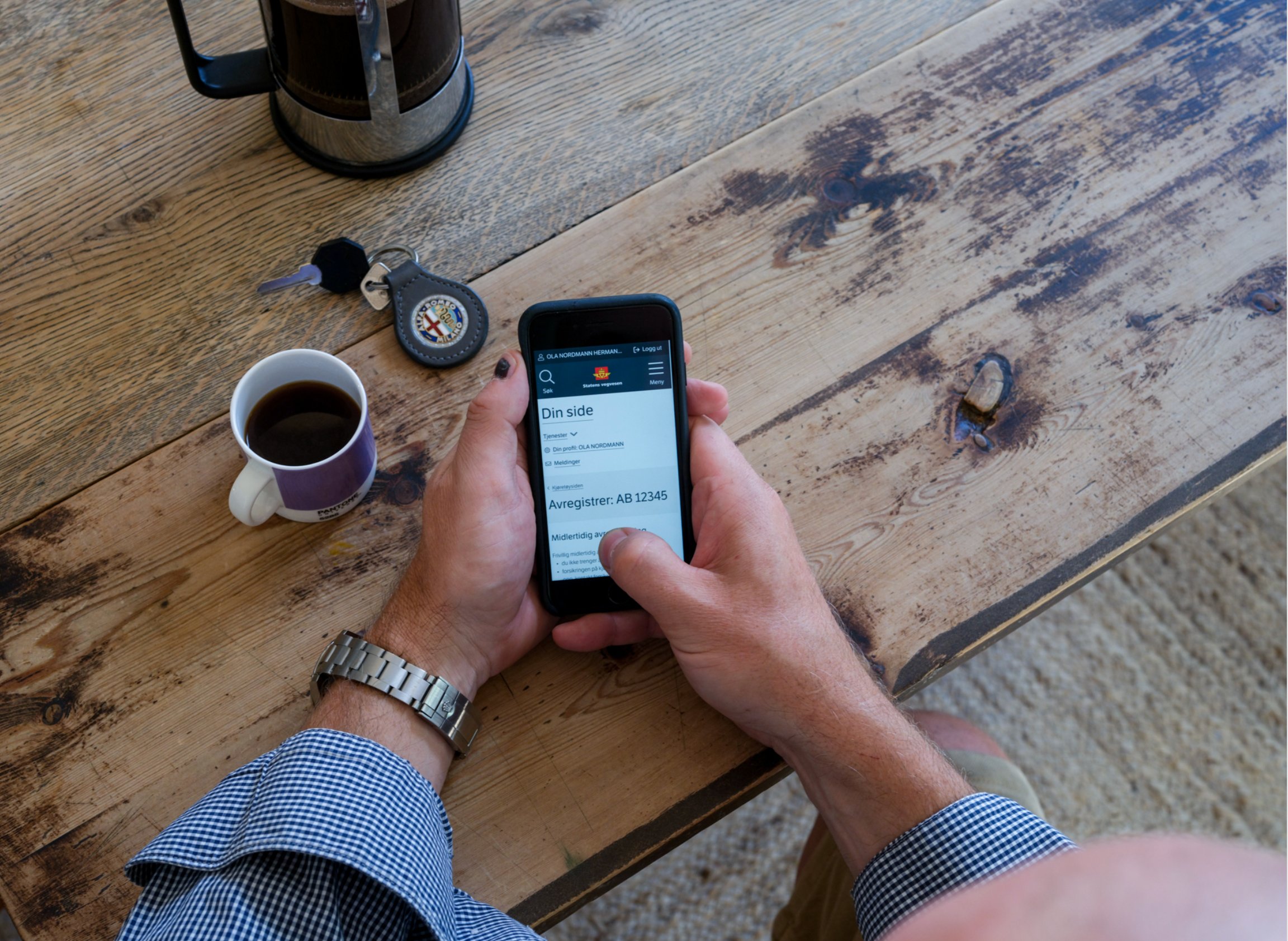 Sett fra perspektivet til en person som sitter ved et bord med en kaffekopp og bilnøkkel på, og personen holder en smarttelefon med Statens vegvesens nettsider åpent. 