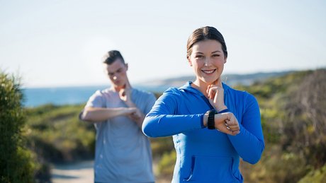 To personer står på en sti ved kysten og sjekker smartklokker, iført treningsklær med hav og vegetasjon i bakgrunnen.