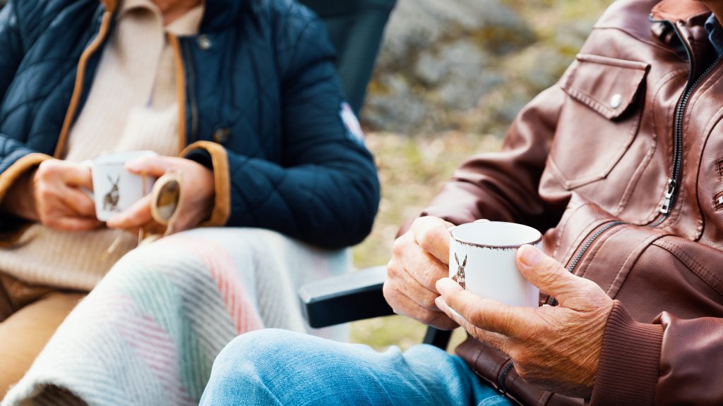 To pensjonister sitter utendørs med hver sin kaffekopp i hendene.