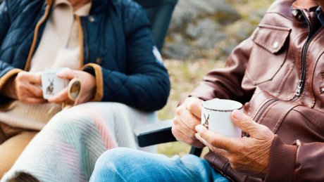 To pensjonister sitter utendørs med hver sin kaffekopp i hendene.