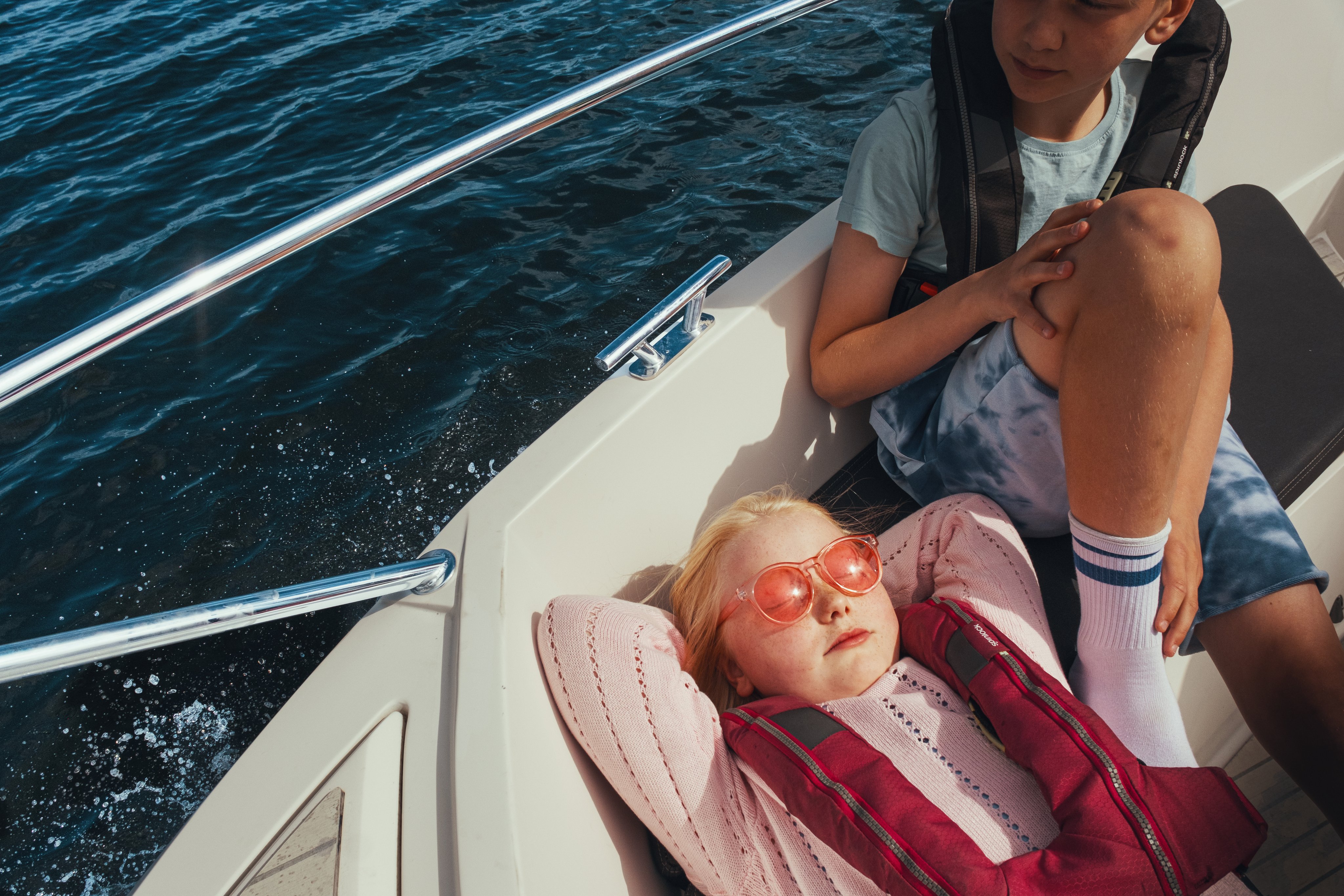 To barn sitter i en båt; én ligger avslappet med redningsvest, mens den andre sitter ved siden av. Vann skimtes ved siden av båten