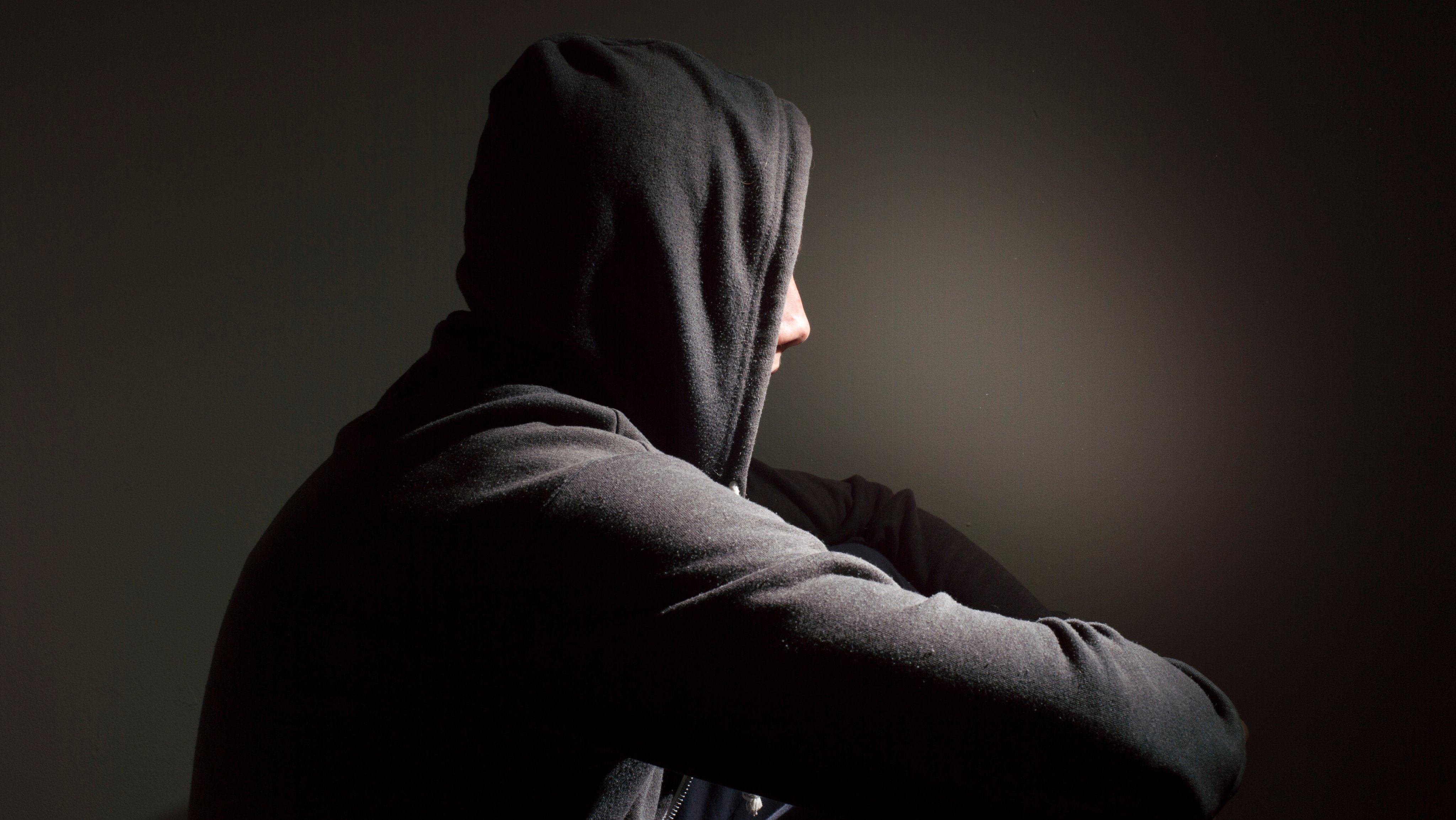 Person med sort hettegenser og hetten på sett fra siden, sittende foran en sort vegg