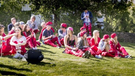 Gruppe ungdommer sitter ute på gresset i solskinn, kledd i røde russeklær og hatter.