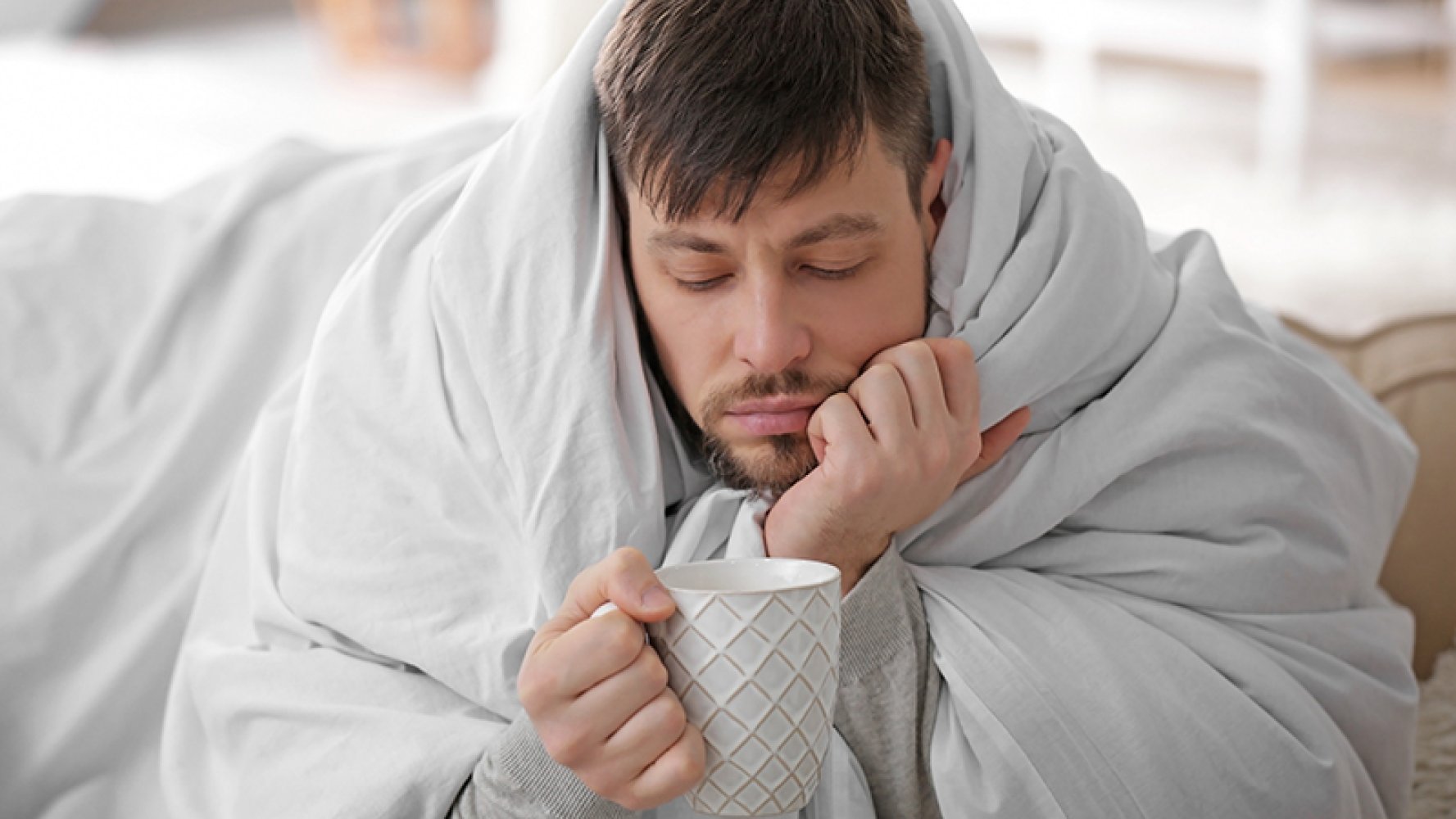 Mann sittende med dynen pakket rundt seg og ser tafatt ned i en hvit tekopp