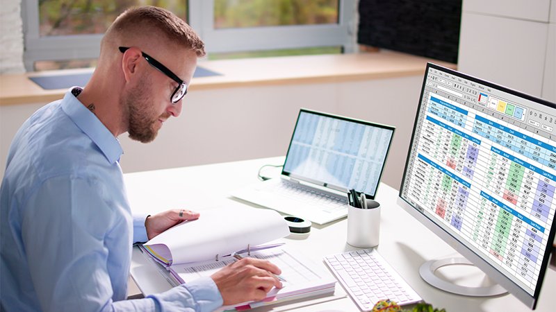 Mann foran to pc-skjermer og en skrivebok, og jobber med regnskap. 