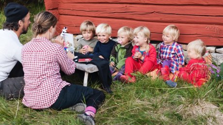 Seks barn sitter foran en rød, laftet vegg.