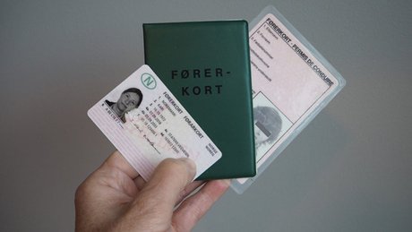 Hånd som holder et norsk førerkort foran en grønn mappe merket ‘FØRERKORT’, med et eldre førerkort i bakgrunnen.