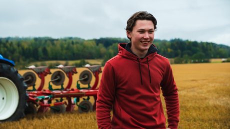 En mann og en kvinne som står med hendene i lommene ute på en gressplen med trær og en rød låve i bakgrunnen.