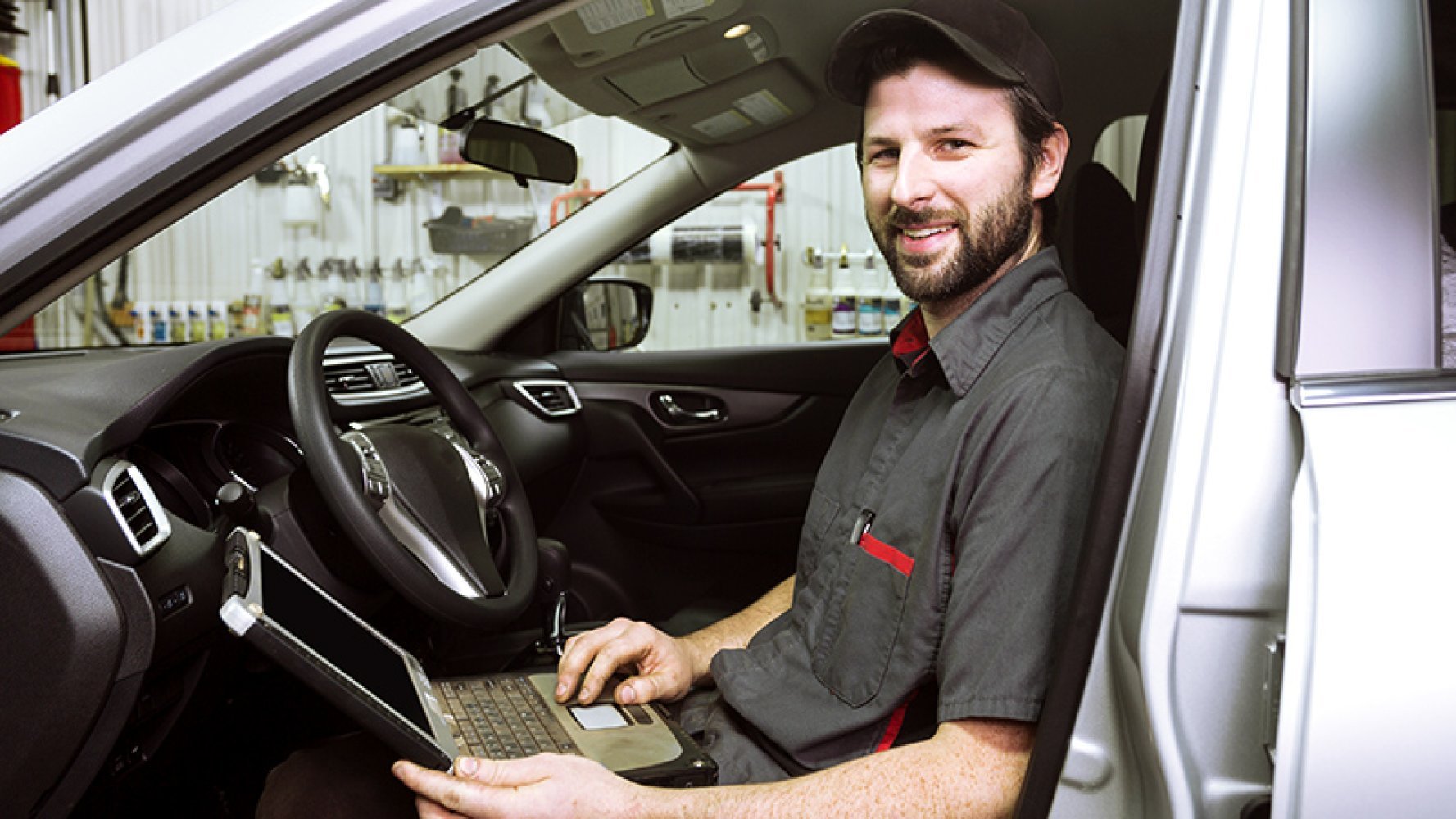 En mann som jobber på verksted sitter i førersetet på en bil og smiler. Han har en laptop i fanget.