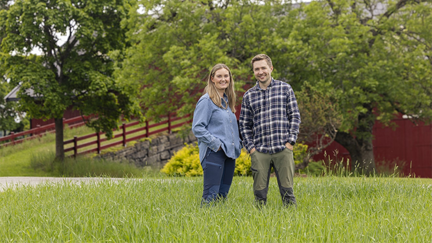 En mann og en kvinne som står med hendene i lommene ute på en gressplen med trær og en rød låve i bakgrunnen.