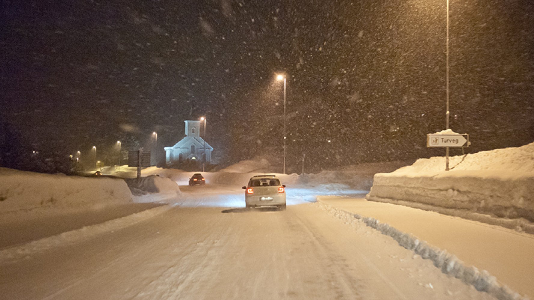 Biler kjører på en snødekt vei i mørket med kraftig snøfall, gatelys og en kirke i bakgrunnen.