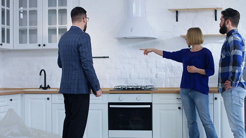 Tre personer står i et moderne kjøkken, mens én person peker mot kjøkkenskapene.