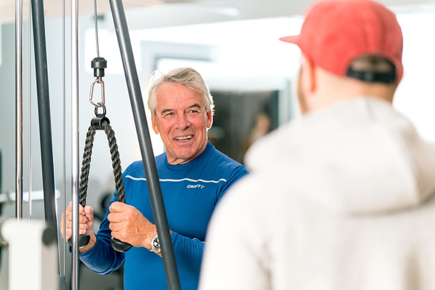 En eldre mann trener med triceps-tau på en kabelmaskin, mens en annen mann med står ved siden av og observerer.