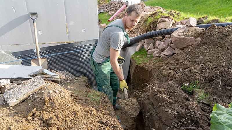 En mann i grønn snekkerbukse graver i hagen med en spade.