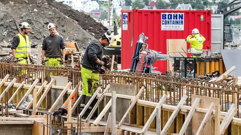 Bygningsarbeidere jobber på en anleggsplass med armeringsjern og forskaling for betong, med en rød container i bakgrunnen og verktøy plassert rundt.