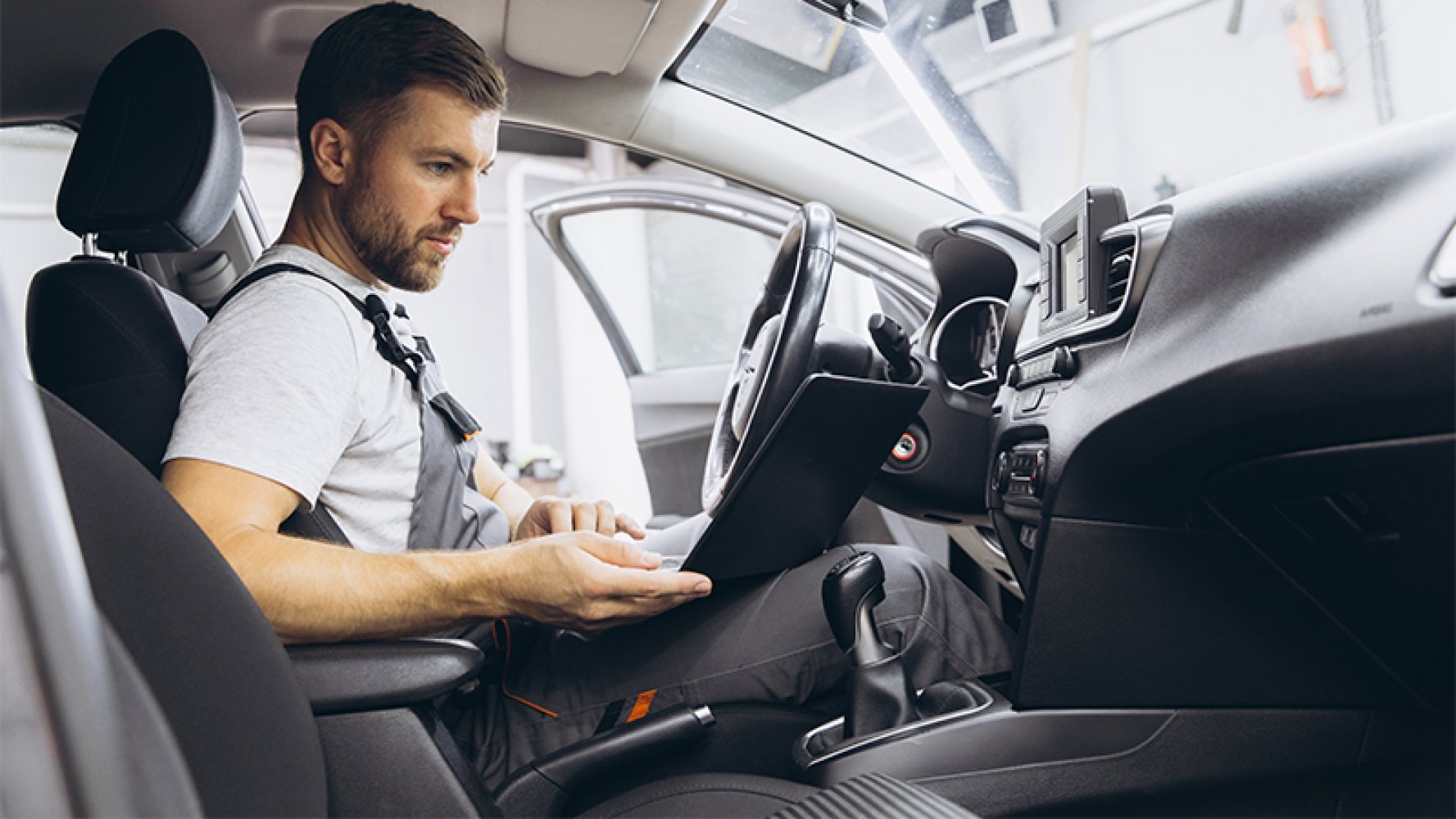 En bilmekaniker sitter i forsetet på en bil med en pc på fanget.