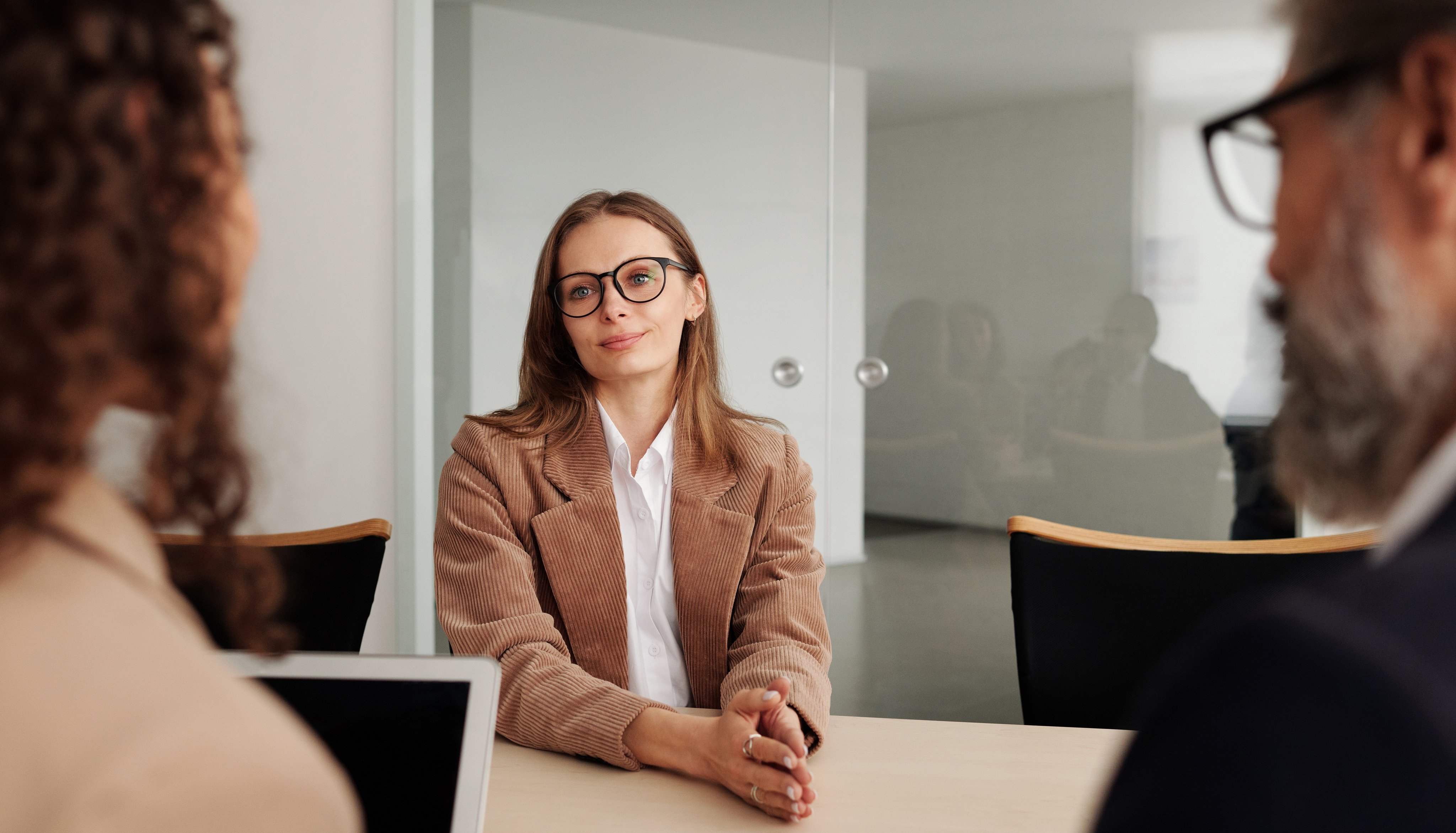 Person sitter ved et møtebord med hendene foldet foran seg, omgitt av to andre personer. I bakgrunnen er en glassvegg og et par personer som reflekteres i glasset.