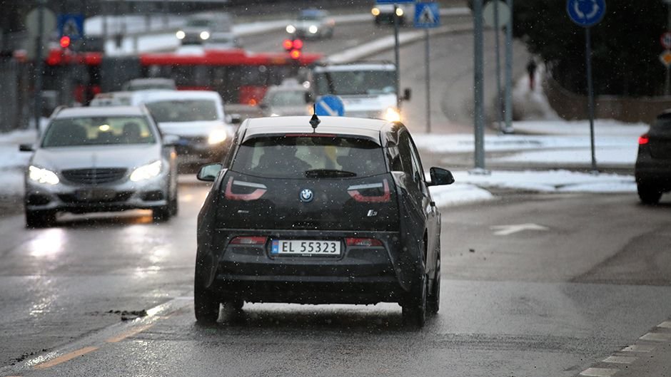 Bil kjører på en våt, snødekt vei i bytrafikk, omgitt av flere biler og busser.
