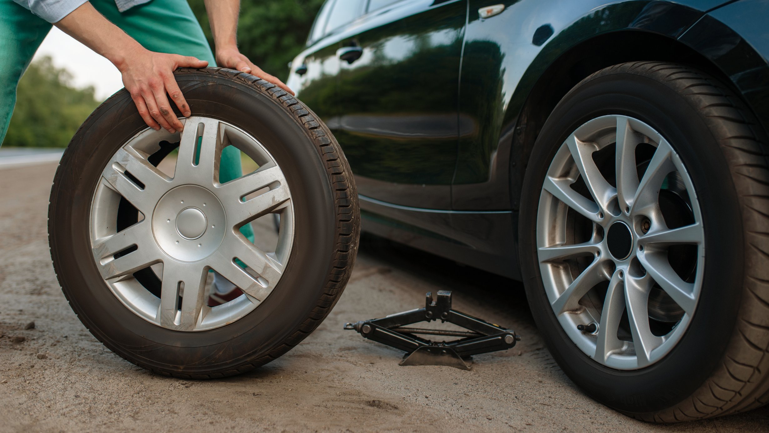 Person bytter bilhjul ved å holde et reservehjul foran en bil med jekk plassert på bakken.