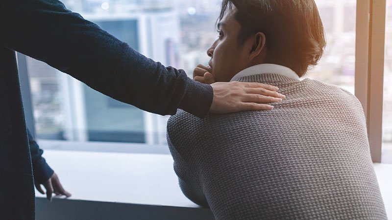 En person legger en trøstende hånd på skulderen til en mann som sitter og ser tankefullt ut av vinduet