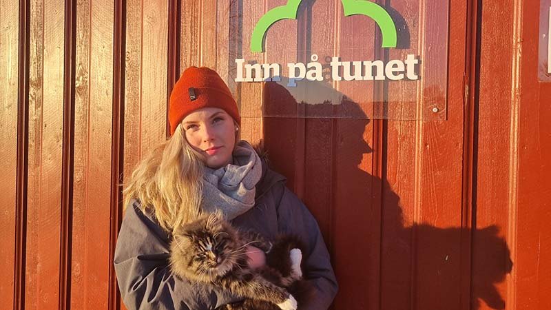 Siv Bjørkedal Åsvestad står foran en rød låvevegg med et skilt hvor det står "inn på tunet".