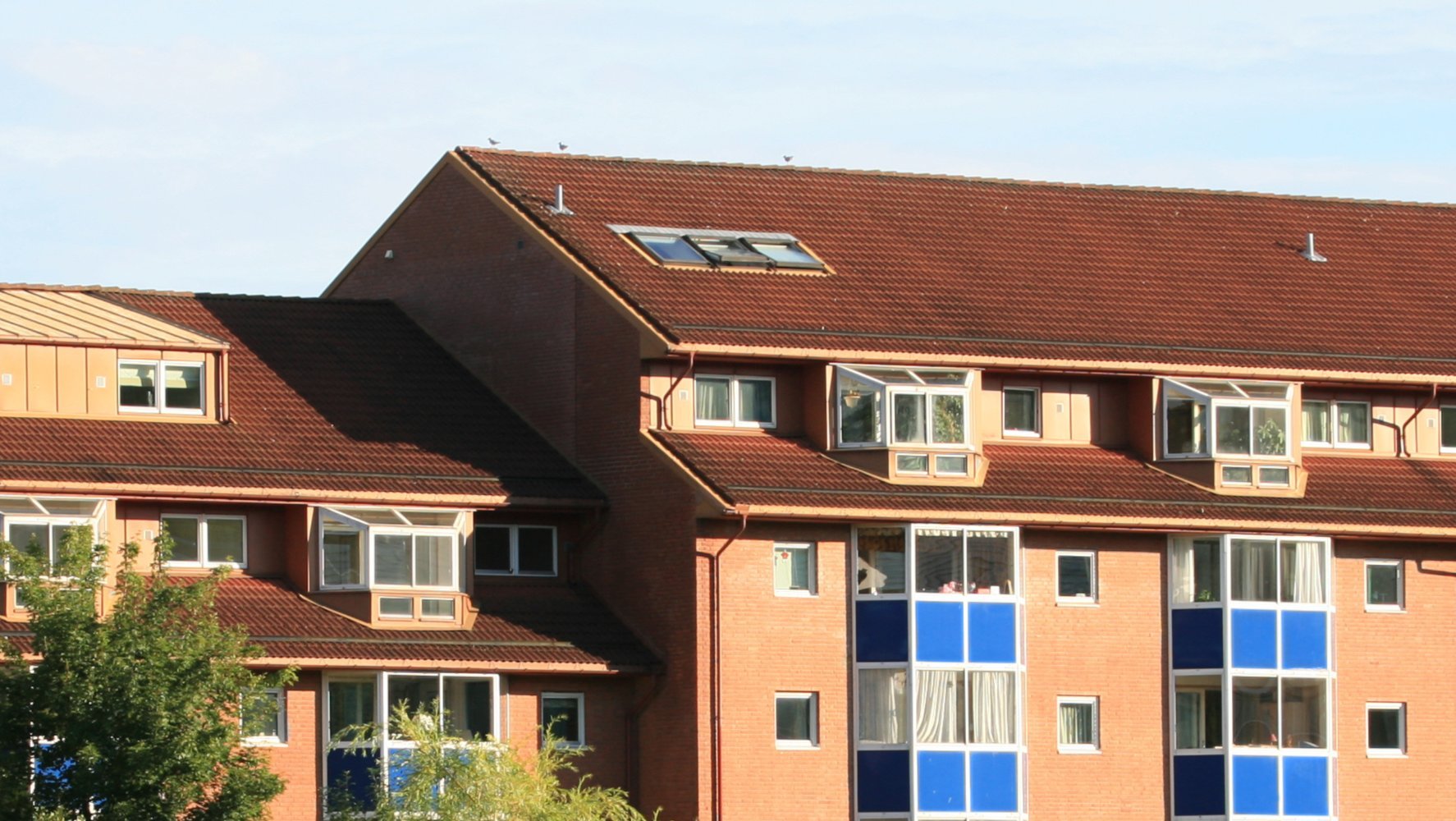 Boligbygg med rødt skråtak, flere vinduer og blå balkongpaneler under klar himmel.