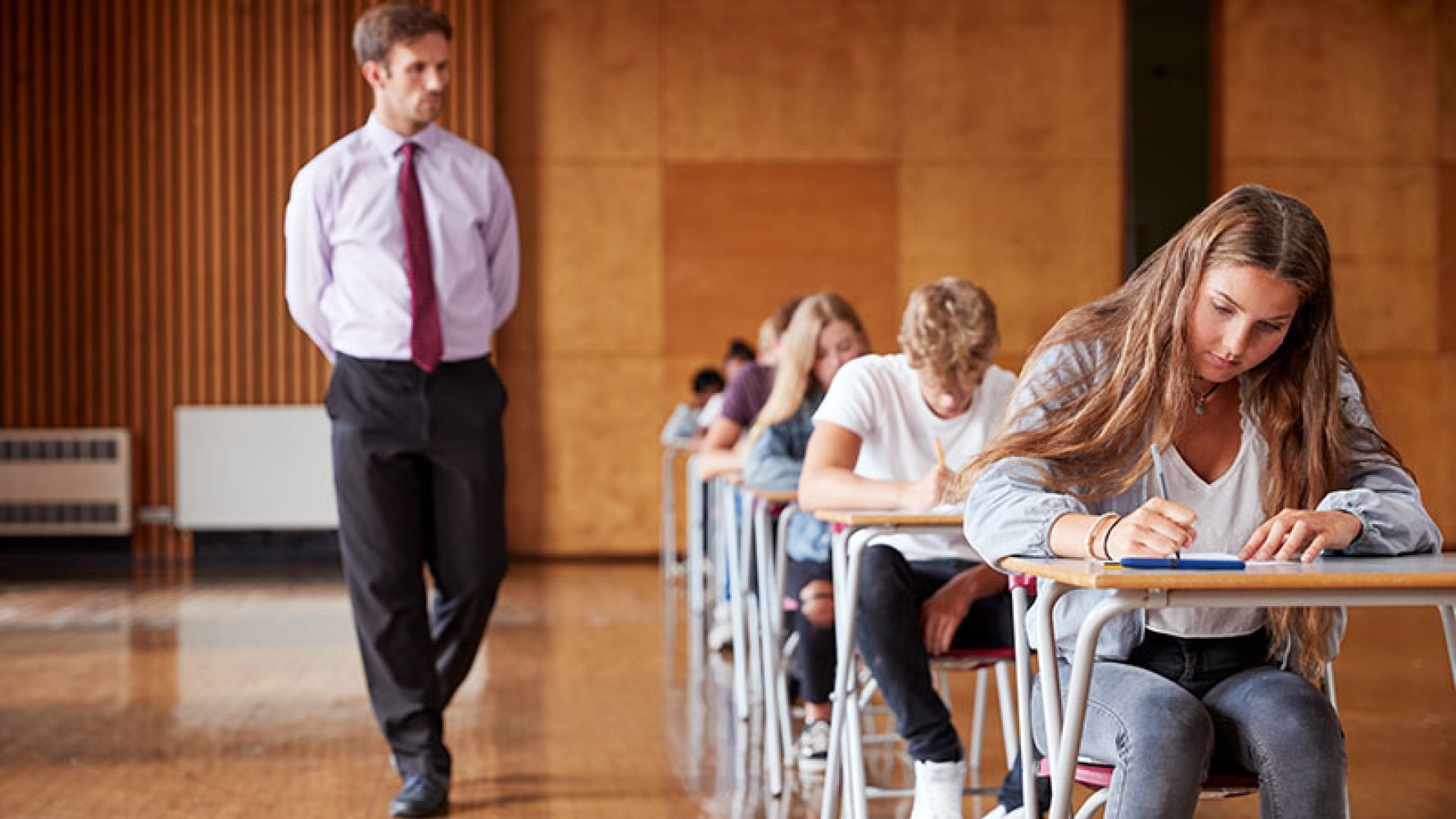 Elever sitter å svarer på en prøve i et stort rom mens en eksamensvakt går forbi.