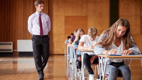 Elever sitter å svarer på en prøve i et stort rom mens en eksamensvakt går forbi.