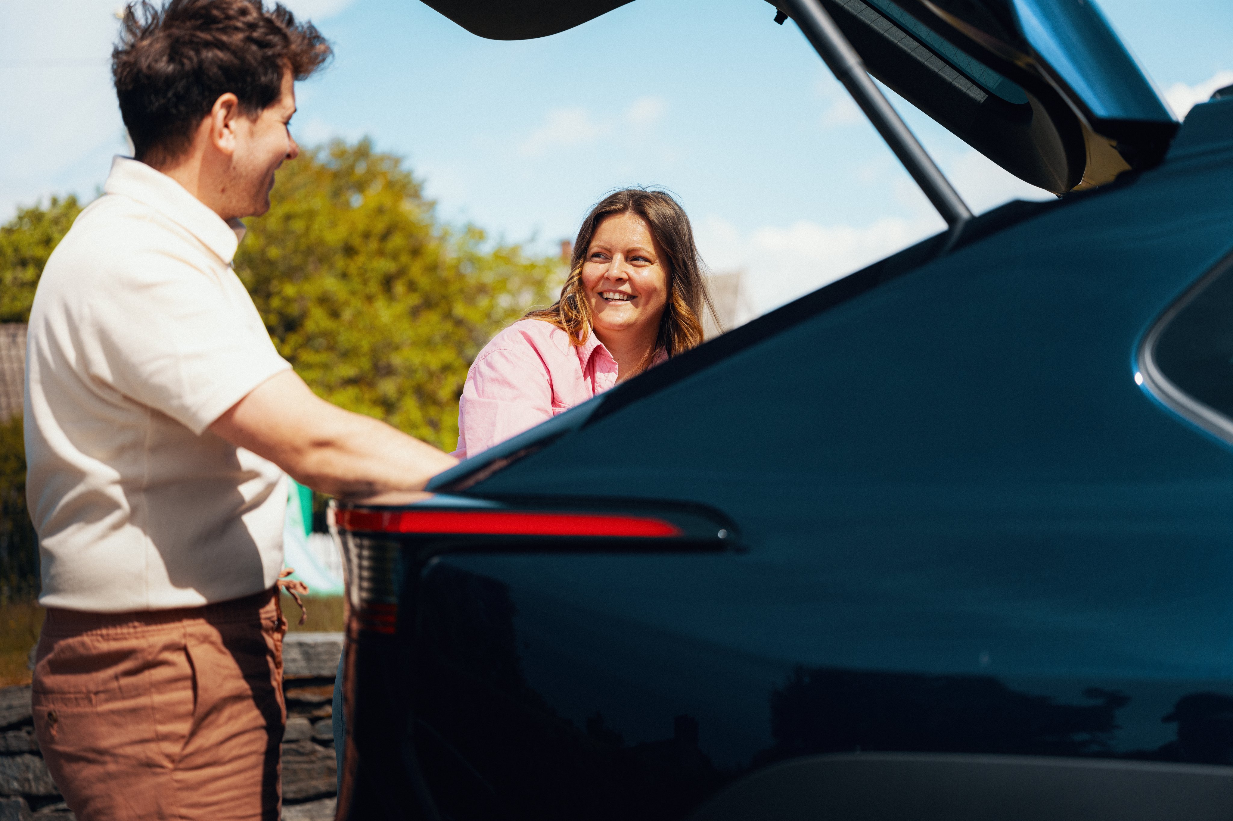 En dame og en mann står ved en bil med åpen bagasjeromsluke, i ferd med å legge inn eller ta ut baggasje.