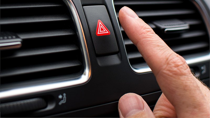 En person er i ferd med å trykke på knappen for nødblinklys i en bil.
