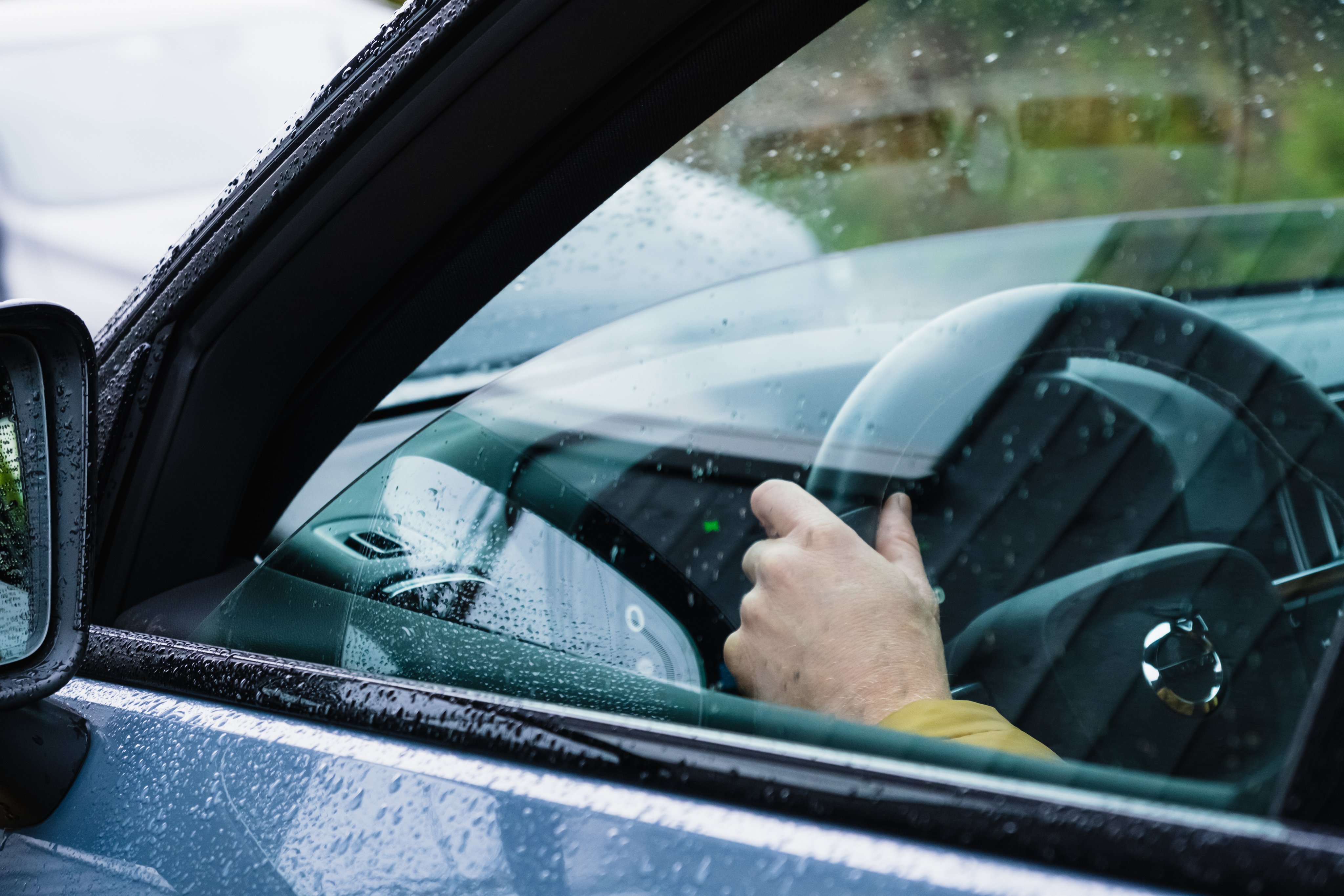 Hånd på rattet sett gjennom en regnvåt bilrute, med sidespeil og dashbord synlig.