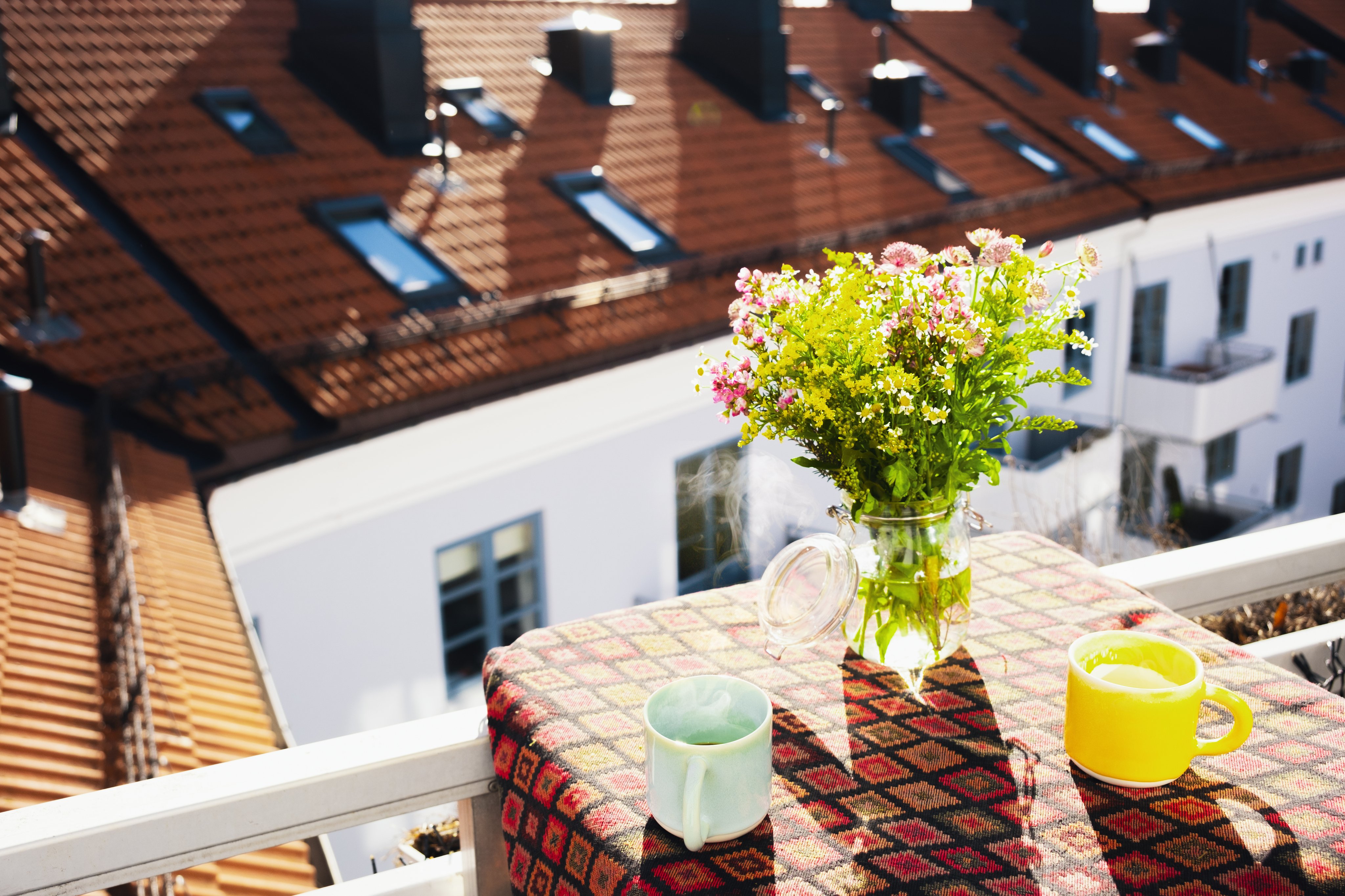 Et lite balkongbord med en duk, to fargerike kopper og en vase med friske blomster i sollys. I bakgrunnen sees tak og fasader fra nabobygg.