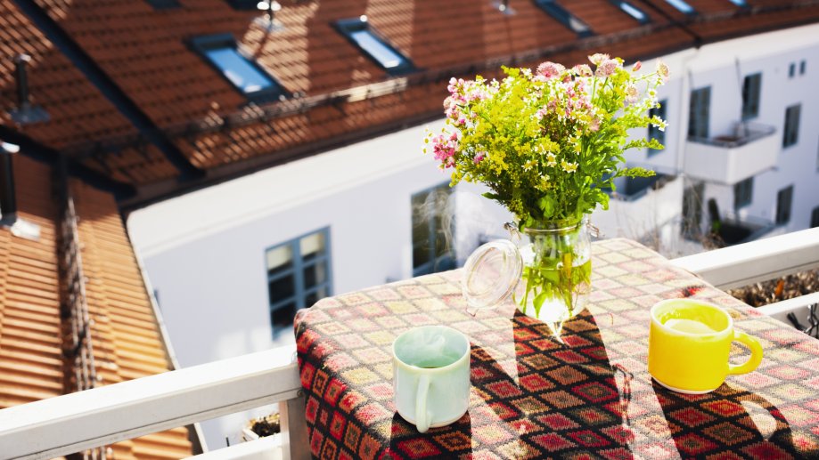 Et lite balkongbord med en duk, to fargerike kopper og en vase med friske blomster i sollys. I bakgrunnen sees tak og fasader fra nabobygg.