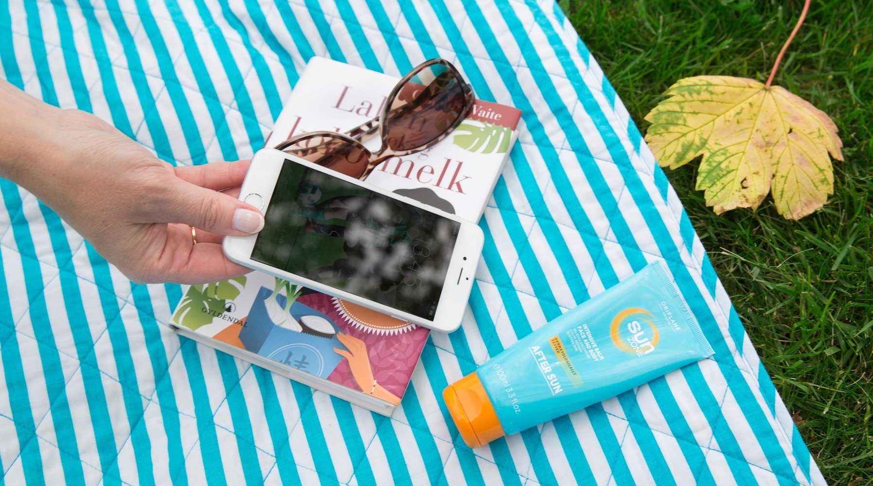 En mobiltelefon med skjermen vendt mot kamera, med teppe, solbriller og solkrem i bakgrunnen.