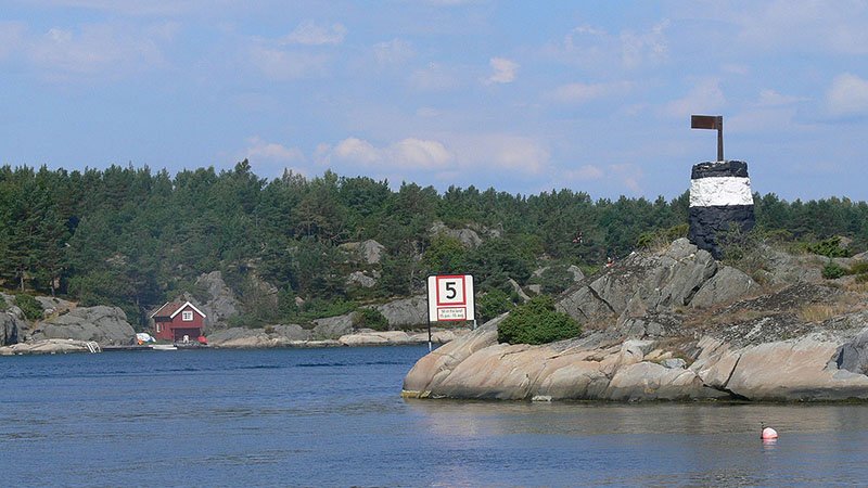 Kystlandskap med steiner, sjømerke og fartsgrenseskilt ved vannet, med skog og en hytte i bakgrunnen.