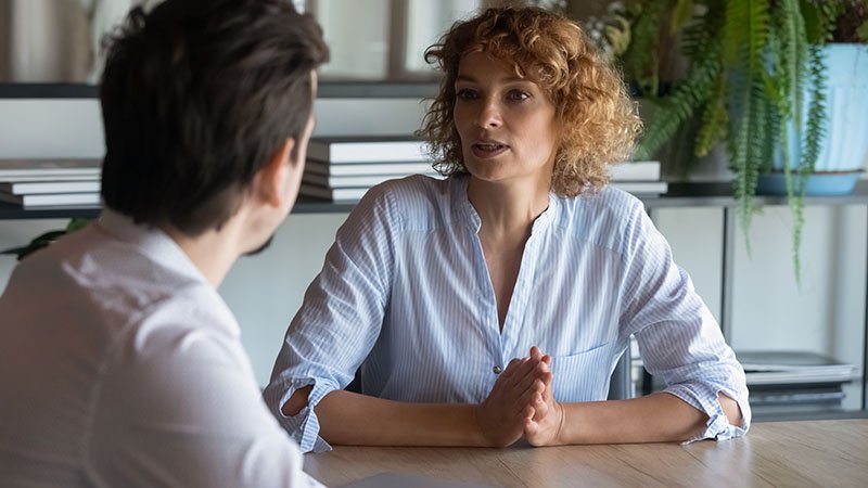 En kvinne sitter ved et bord og snakker med en mann i et lyst kontormiljø.