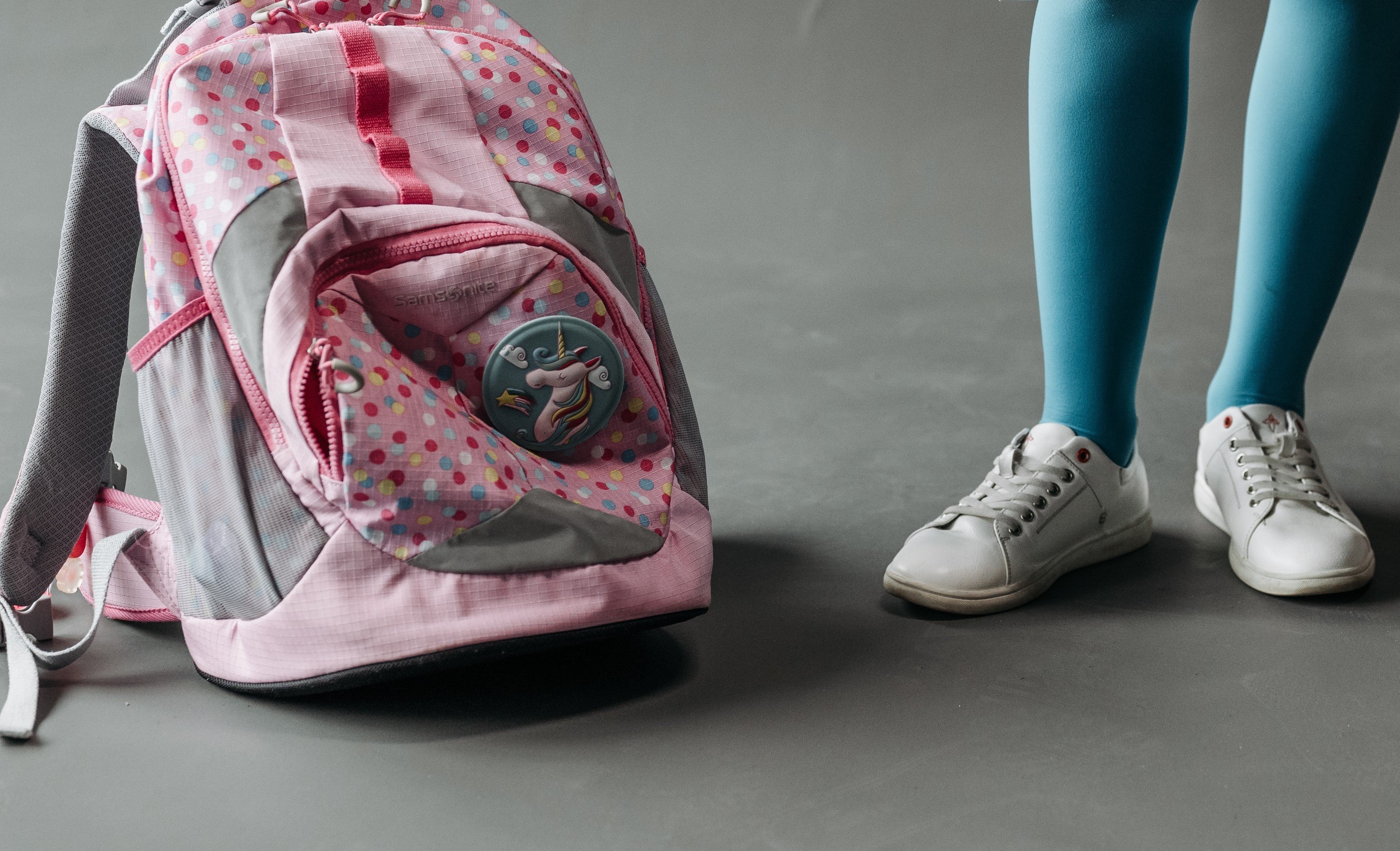 Ryggsekk med fargerike prikker og en enhjørning-merke står på gulvet ved siden av beinene til et barn.