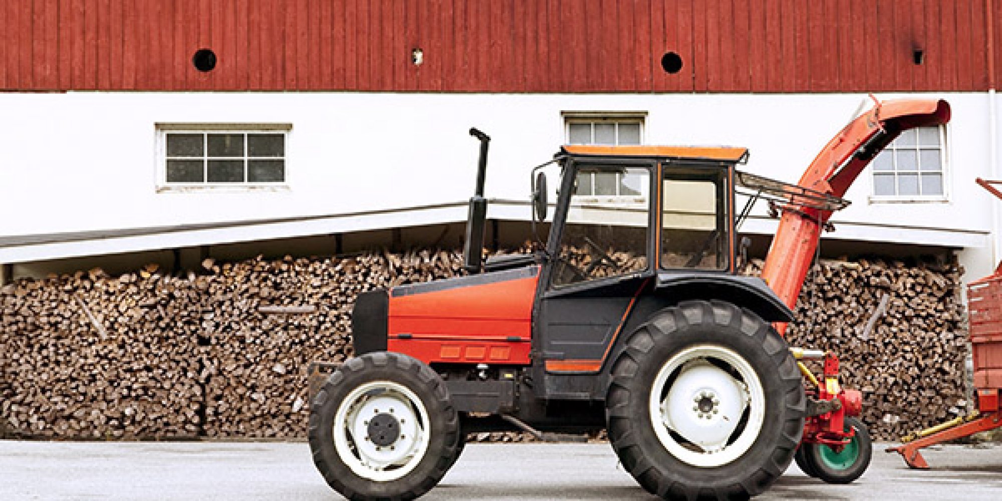 Eldre traktor stående foran en låve, med store mengder ved stablet langs veggen under et tak.