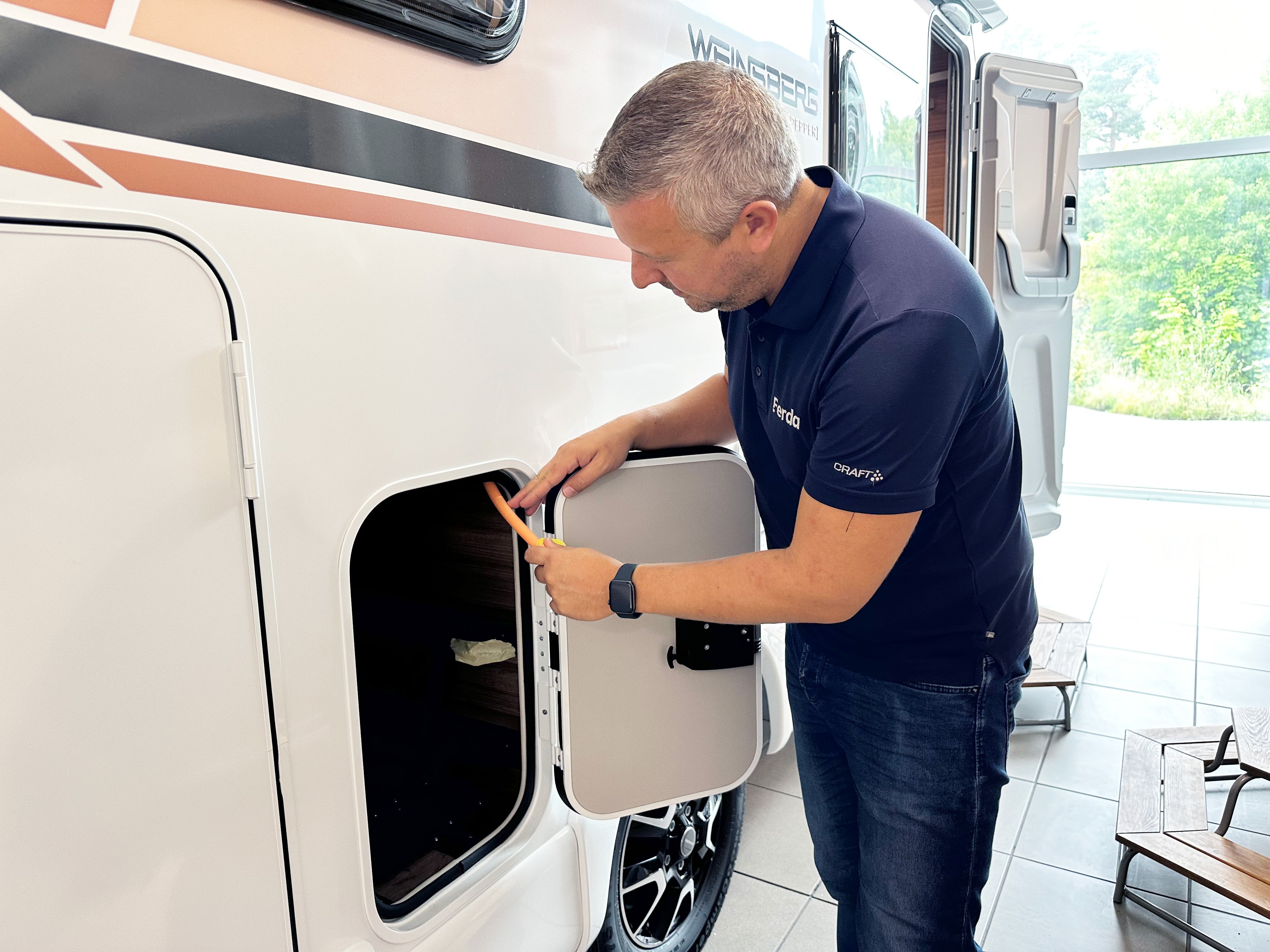 Mann som åpner en luke på utsiden av en bobil og undersøker rommet med et verktøy.