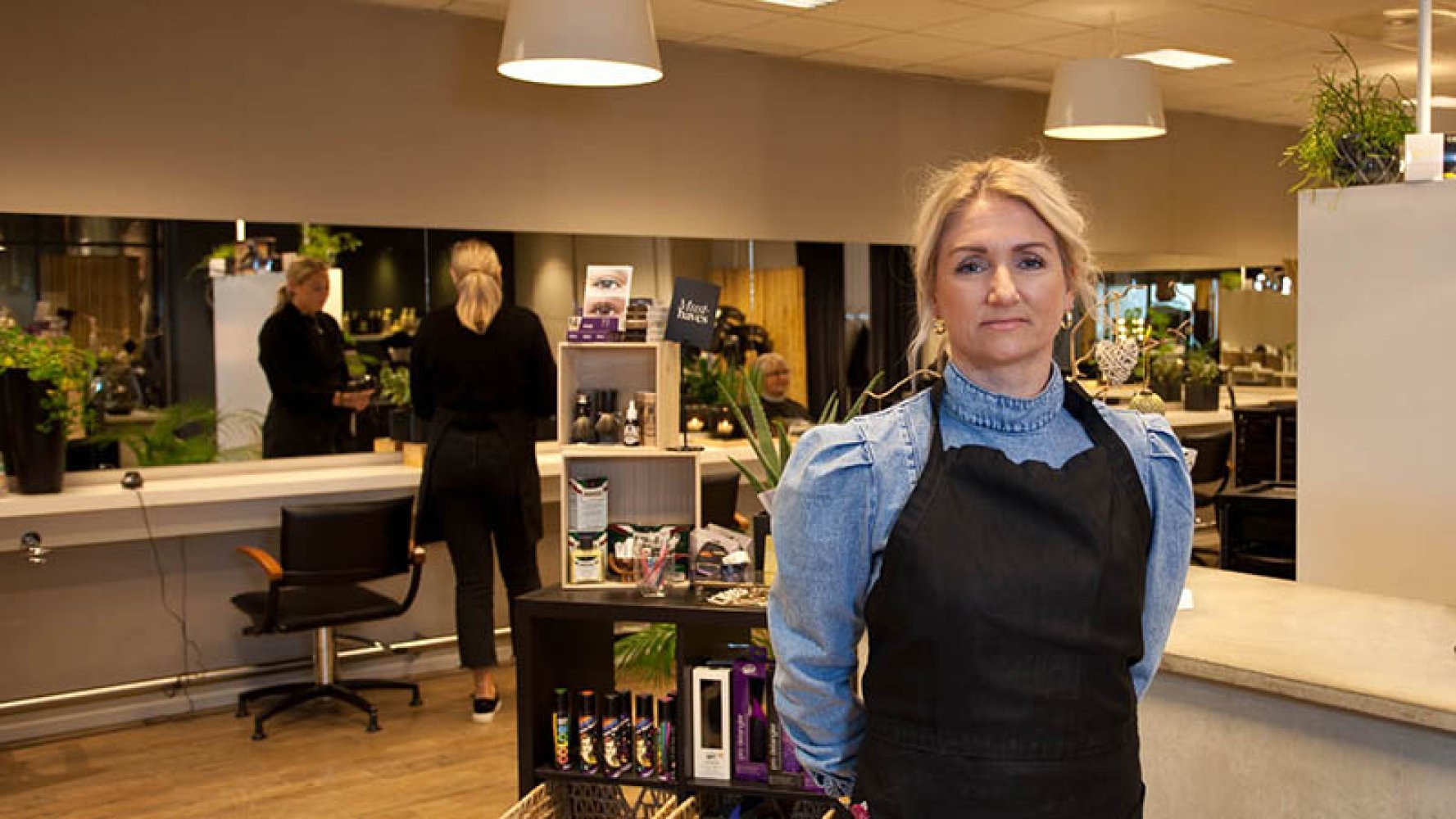 Dame står foran i bildet med hendene på ryggen i frisørsalong med salongen i bakgrunnen.