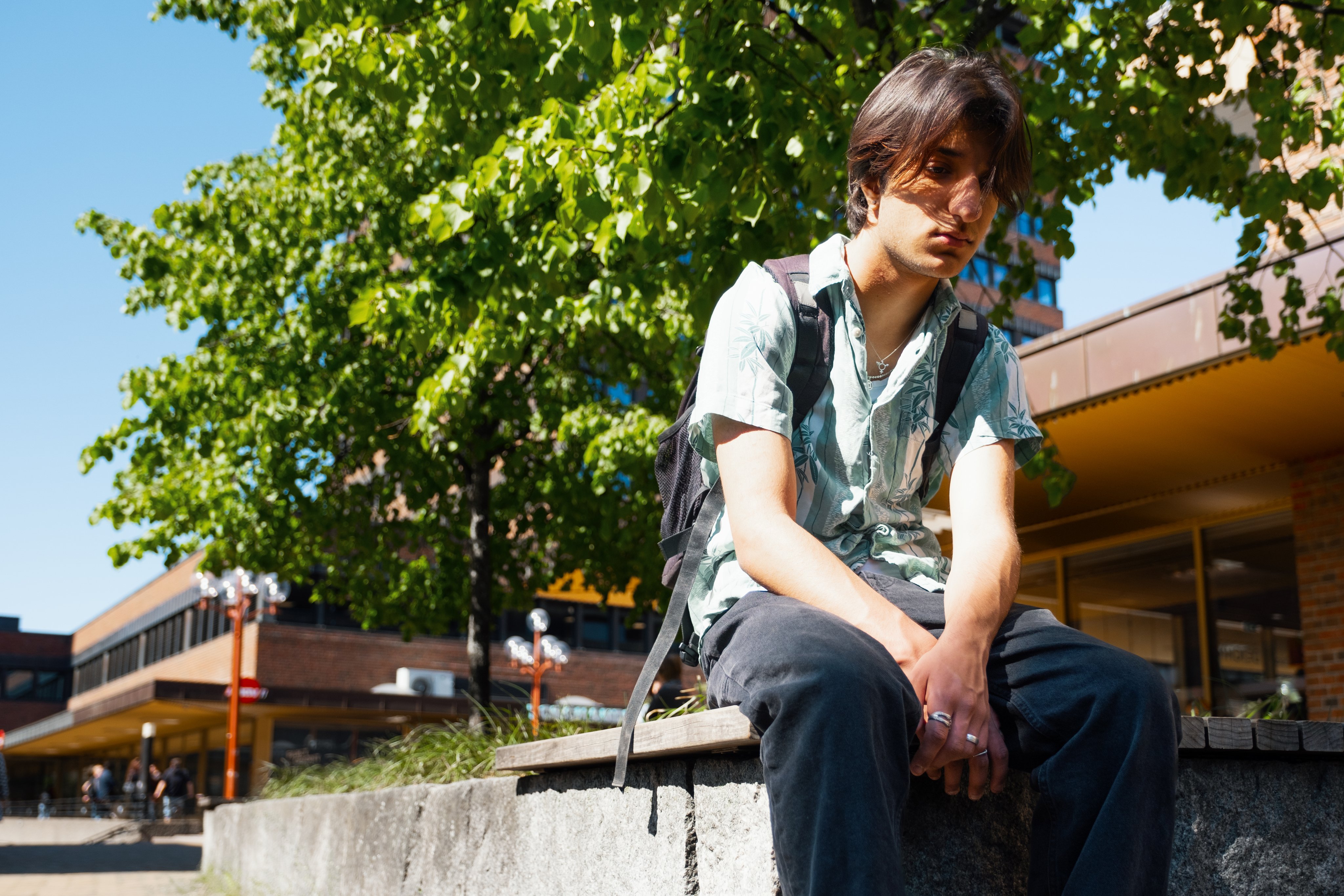 En ung gutt sitter utenfor skolen med sekk på ryggen og ser trist ut.