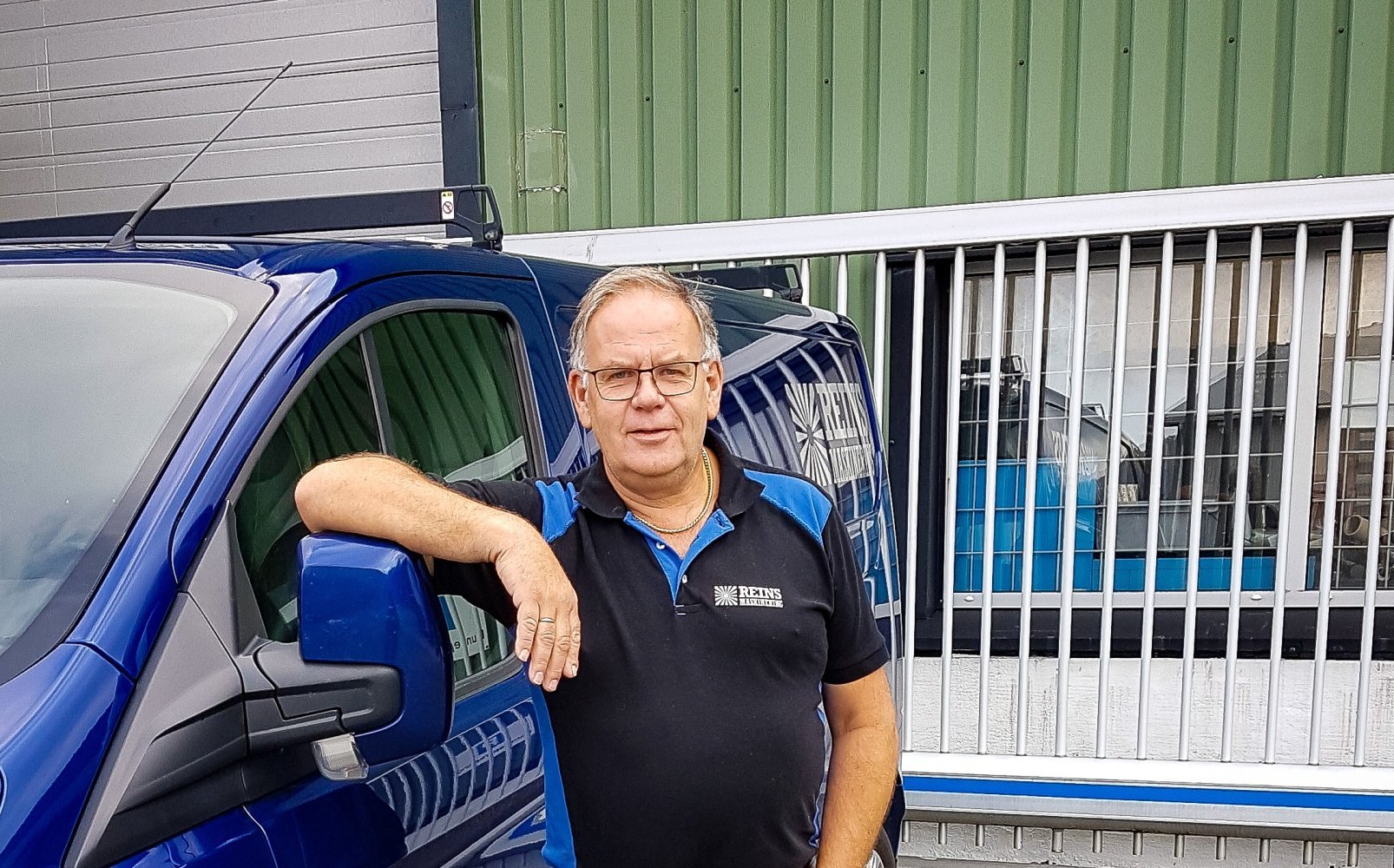 En mann står lent mot en blå firmabil utenfor et industribygg, iført svart og blå arbeidsskjorte med firmalogo.