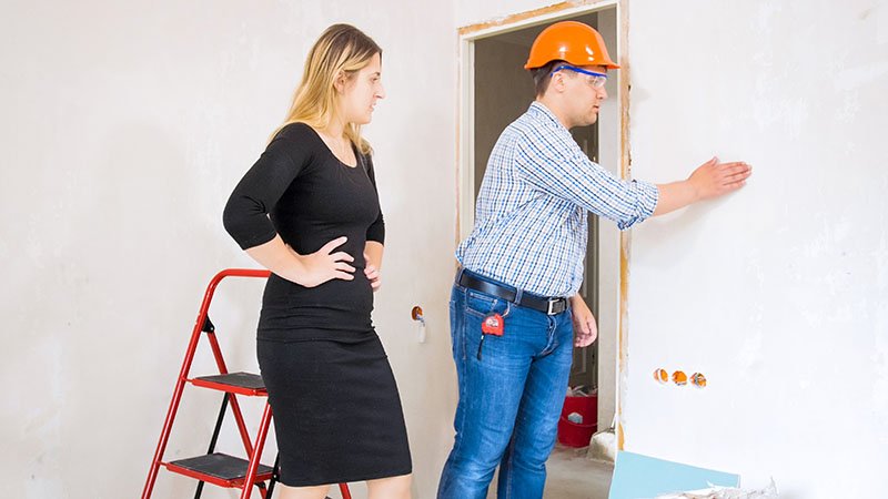 Personer inspiserer en hvit vegg i en bolig under bygging, med en rød stige og verktøy synlig.