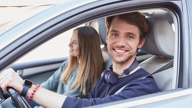 En mann sitter i førersetet i bilen med hodetelefoner rundt halsen og smiler. I bakgrunnen ser vi en jente i passasjersetet.