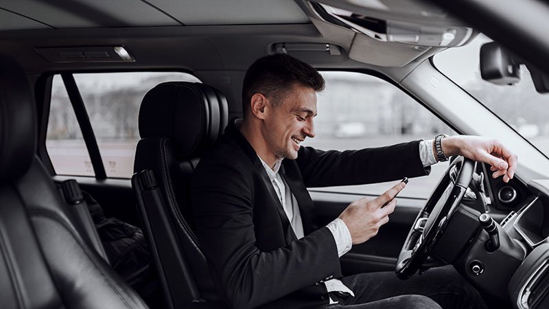 En ung mann sitter i førersetet i en bil og smiler og ser på mobilen han holder i hånda.