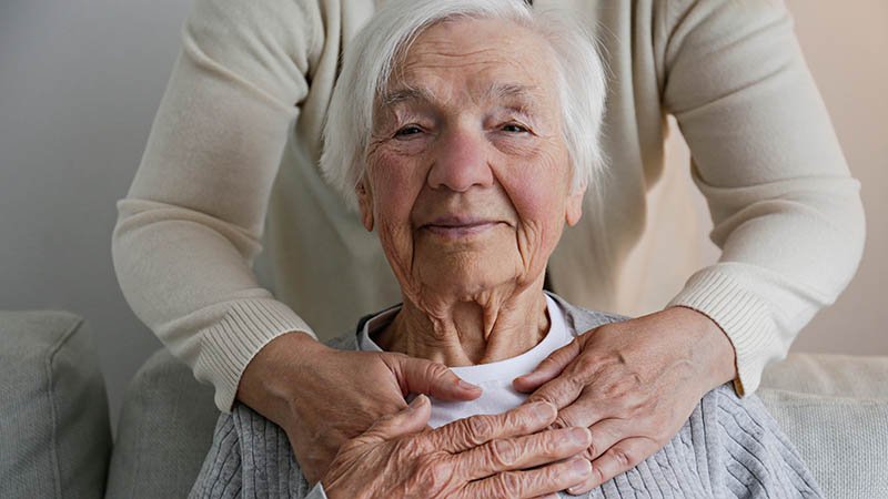 Person holder armene rundt skuldrene til en eldre person som sitter i en stol.